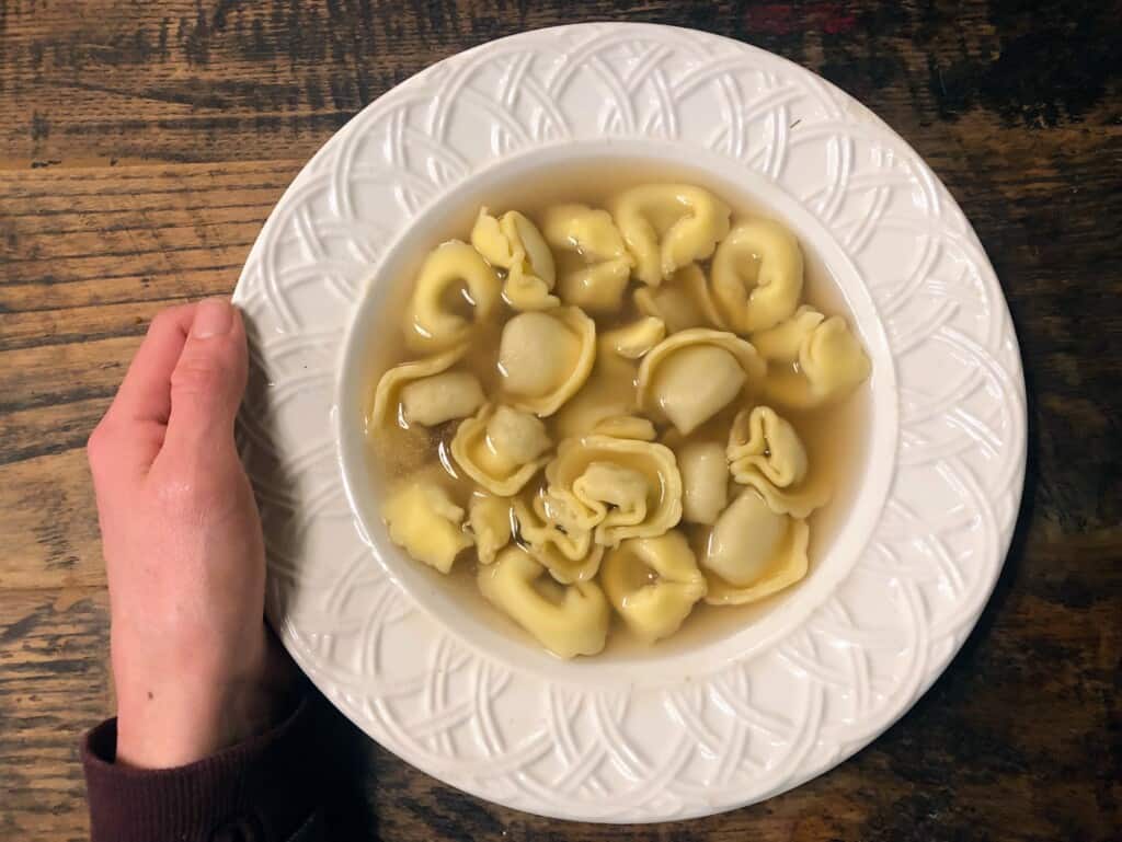 white ceramic shallow bowl filled with tortellini in brodo on a wooden board. a person's hand holding on left side of the bowl from birds eye view.