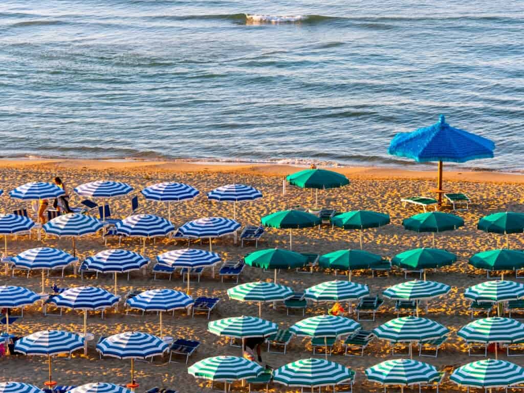 a beach with green and striped umbrellas covering it with sea in background from birds eye view