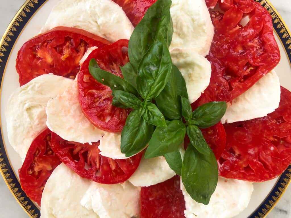 top view of a caprese salad on a white plate with gold trip garnished with a big bunch of basil in the center of the tomatoes and mozzarella.