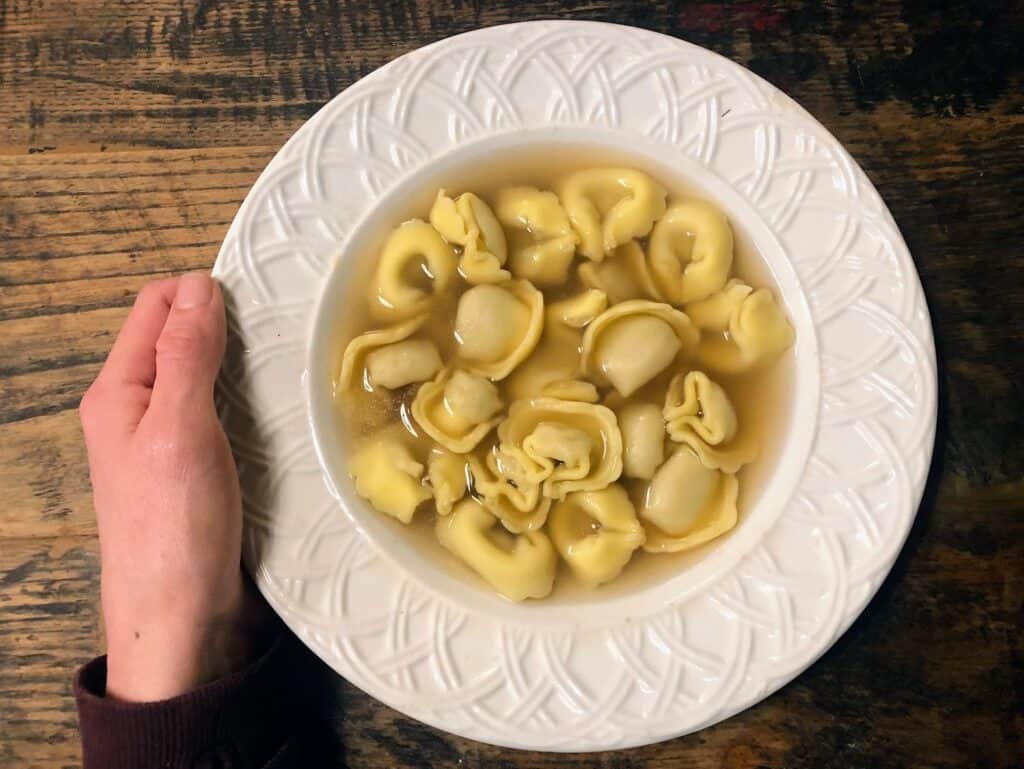 Hand holding a shallow white bowl filled with tortellini in brodo, on top of a wooden table.
