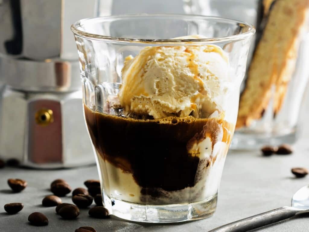 small clear glass with a scoop of vanilla ice cream and a shot of espresso in it with coffee beans on ground and coffee moka pot in background