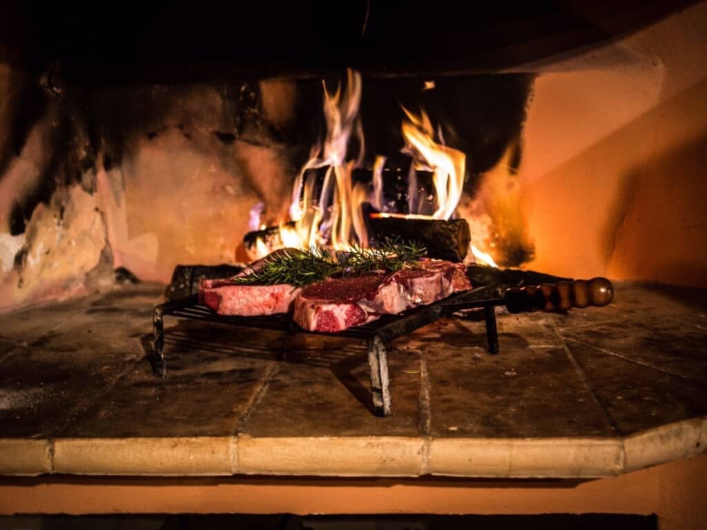 a bistecca fiorentina being grilled on an open fire indoors with a sprig of rosemary.
