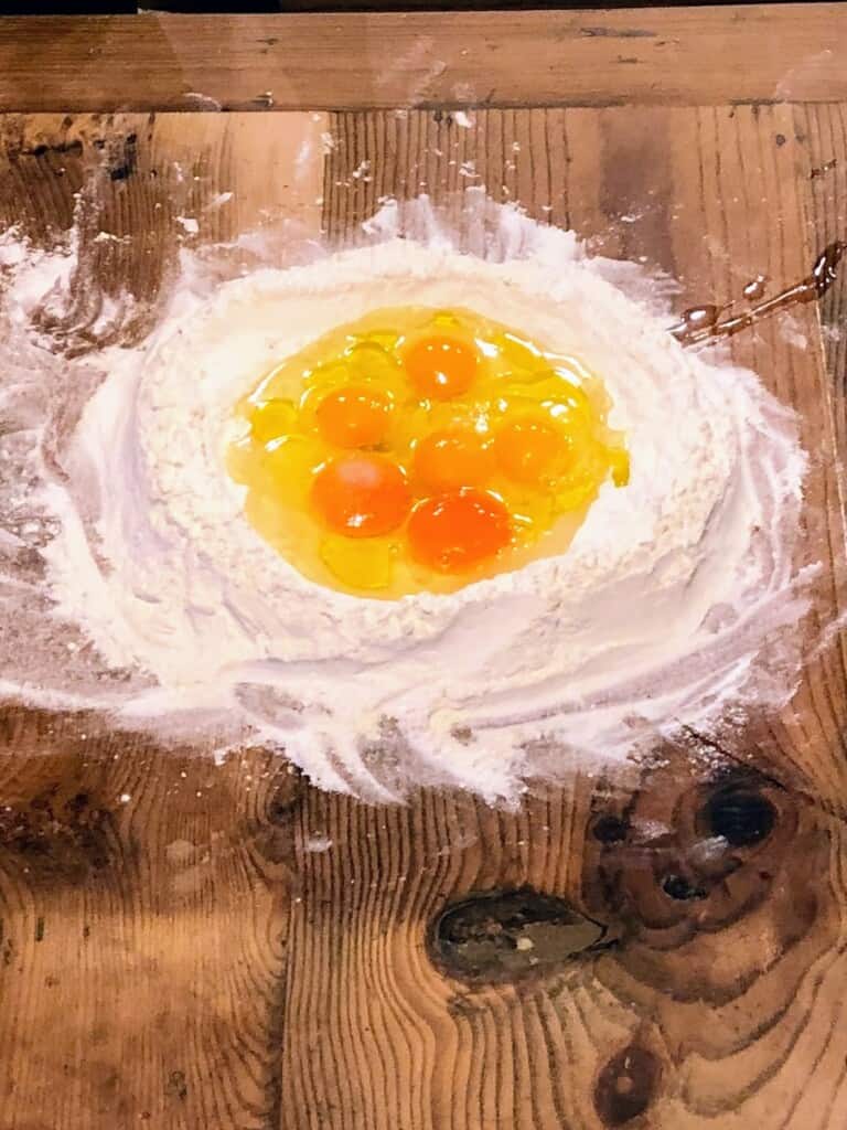 close up of a well of flour with 6 eggs cracked into the center on a wooden board