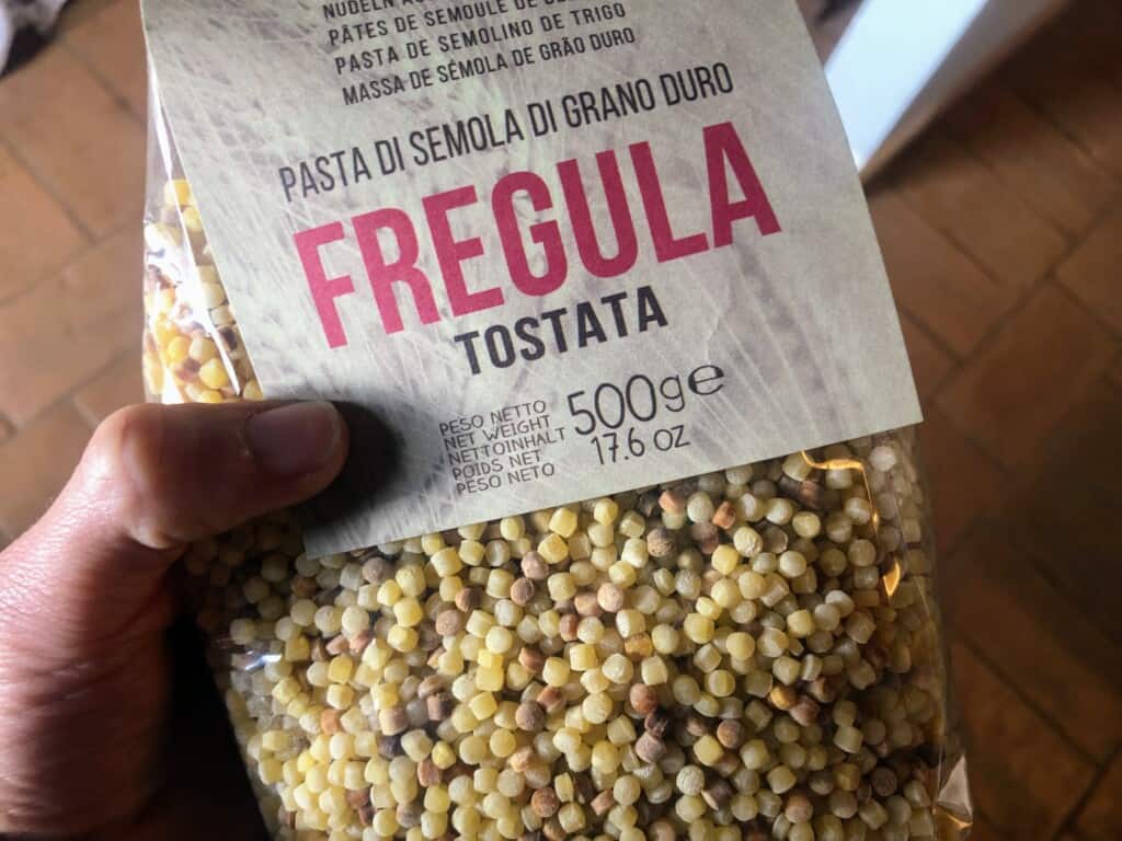 close up of hand holding a package of toasted fregola pasta on wooden background