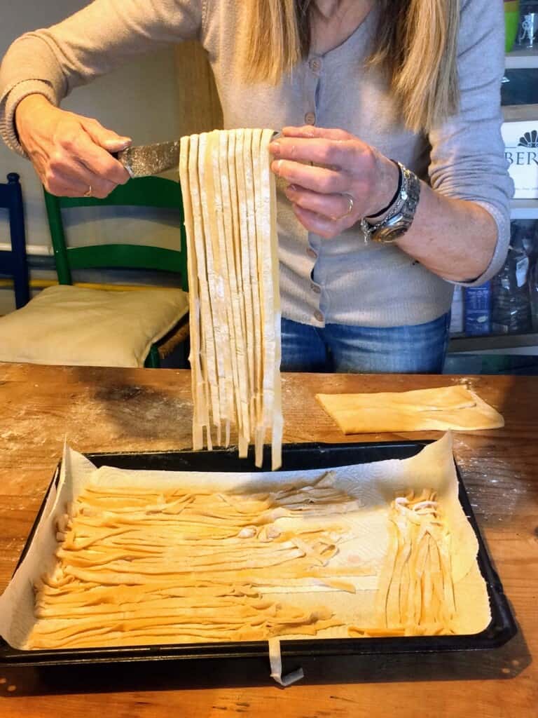 woman making homemade tagliatelle with a pan of pasta already prepared on wooden board in foreground