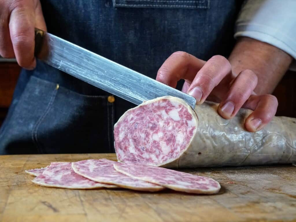 man holding a sausage and cutting it thinly with a large knife on wooden cutting board