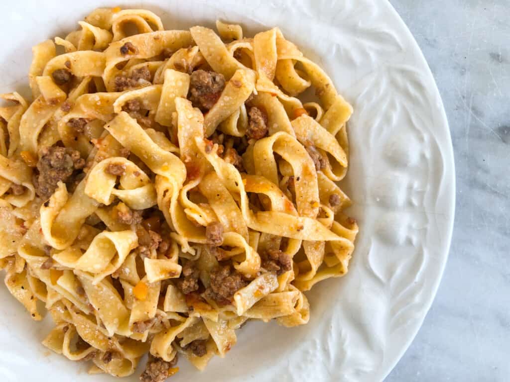 close up of white plate with tagliatelle dressed in ragu sauce on a white marble background showing on right hand side of picture.