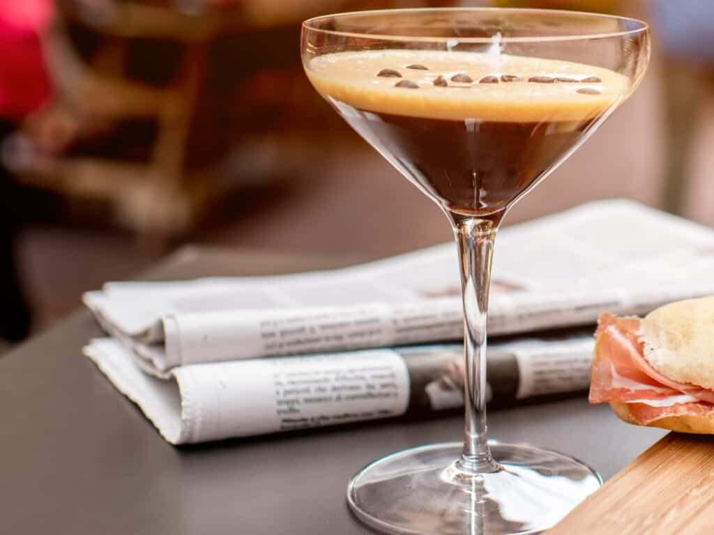 shakerato drink on a table with a newspaper in the background and the corner of a sandwich from side view