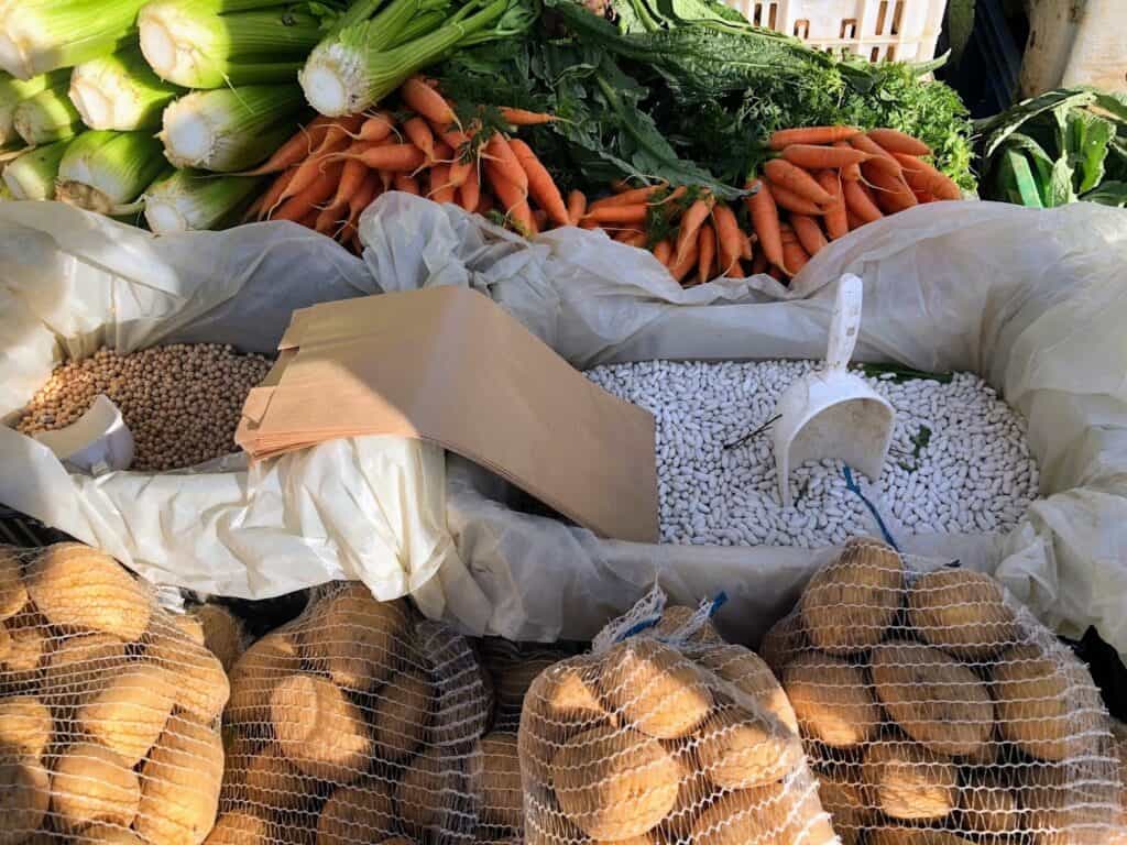 side view of several market items for sale including potatoes on the bottom, white beans in bins in middle and carrot and celery on top.
