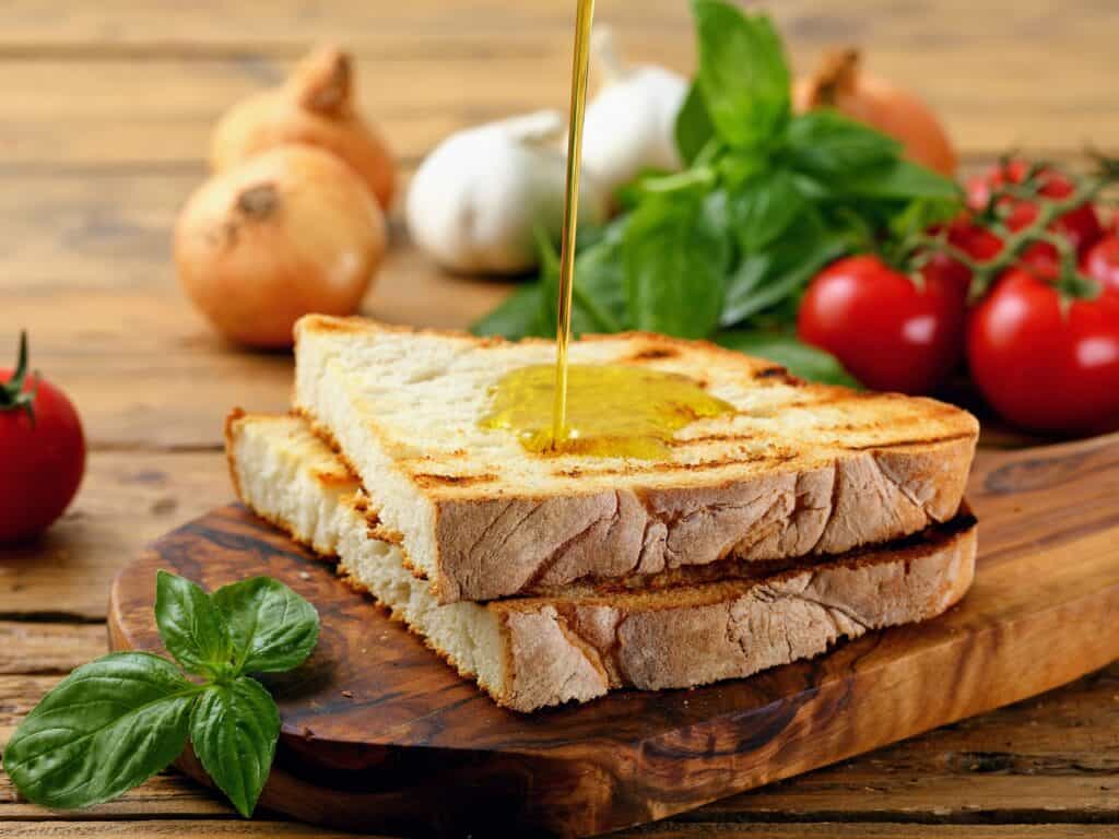 oil being poured on two slices of bread. surrounded by basil, onions, garlice, adn tomatoes.