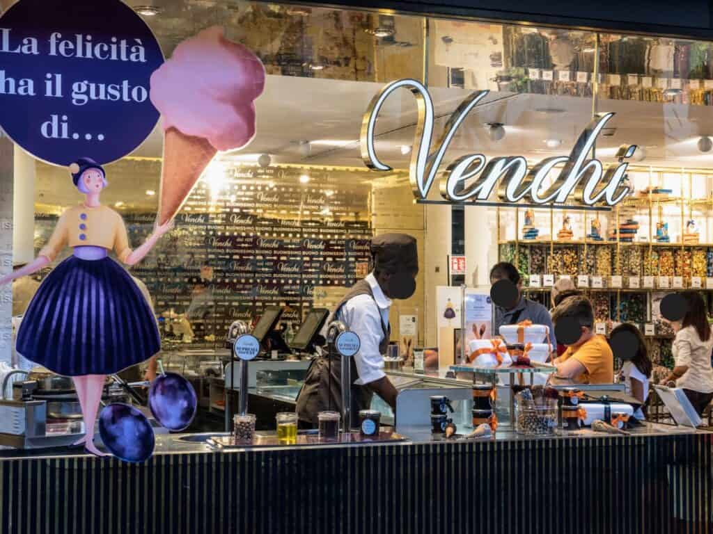 Storefront window of Venchi chocolate.  You can see a woman scooping gelato and people waiting in line and browsing the shelves of Italian chocolates.