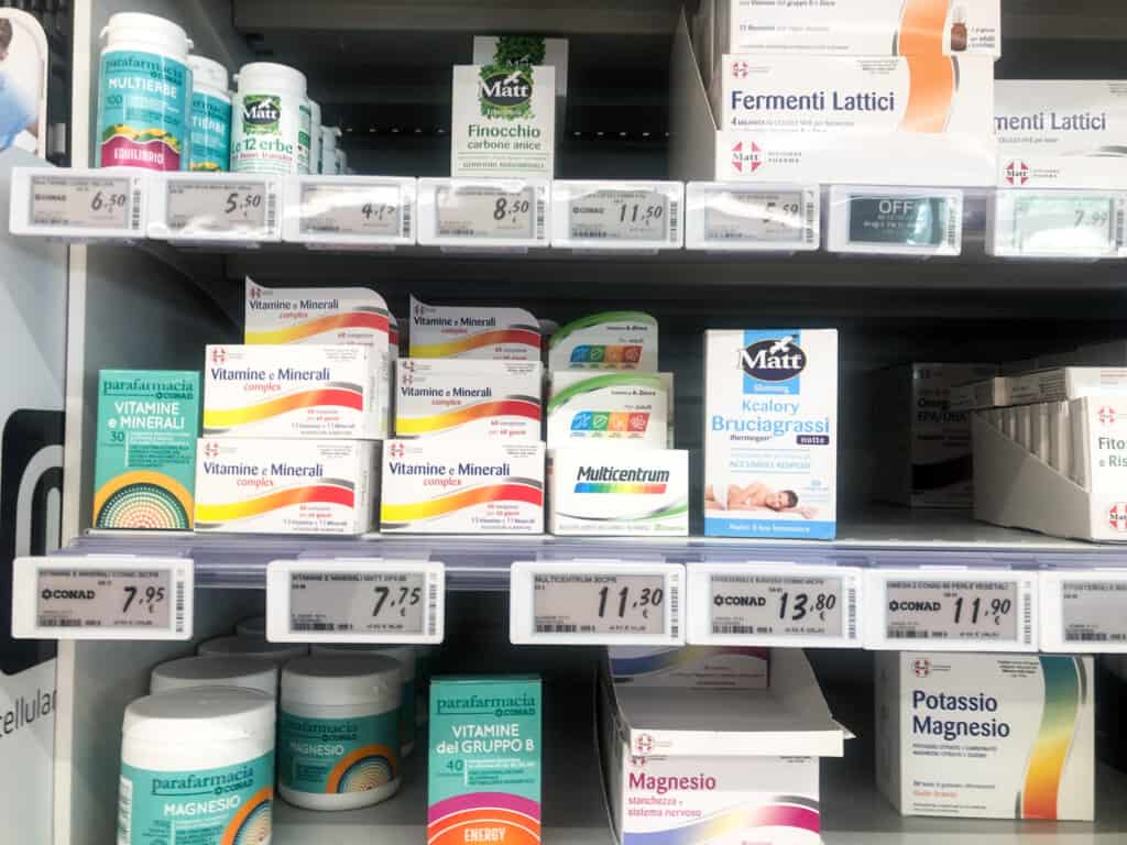 close up of several grey shelving at a grocery store from frontal view with various health products like vitamins on them for sale with price tags underneath
