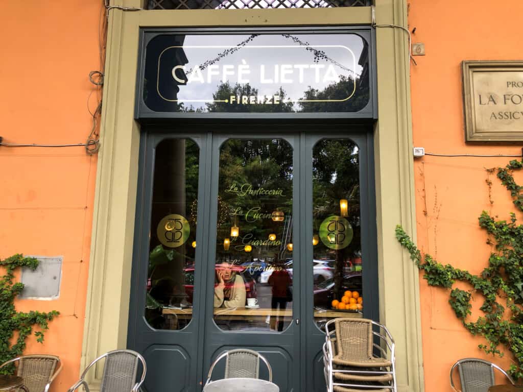 Front door of Caffè Lietta in Florence, Italy. You can see someone sitting at a table inside. There are chairs stacked outside and ivy growing on the wall.
