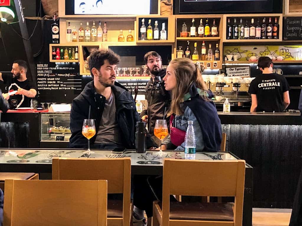 Man and woman sit at bar in Italy. There are drinks in front of them. In the background you can see the bar and bartenders and bottles on the shelves.