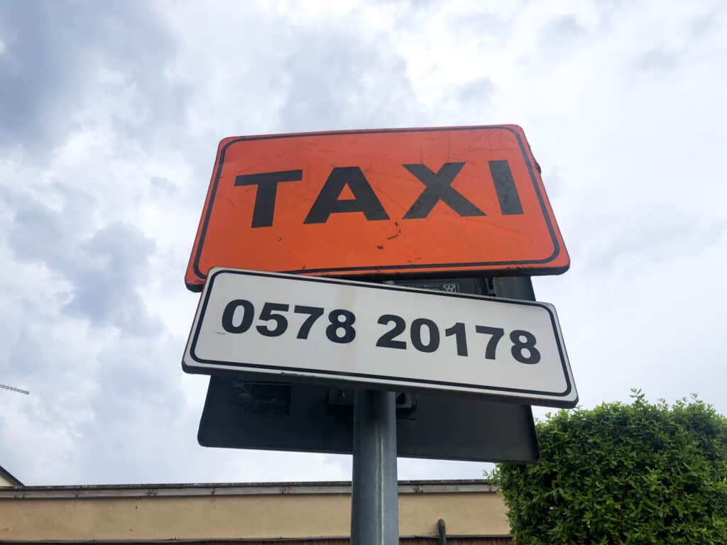 Orange taxi sign with white sign of phone number just below it. You can see sky and the top of a bush and building in background.