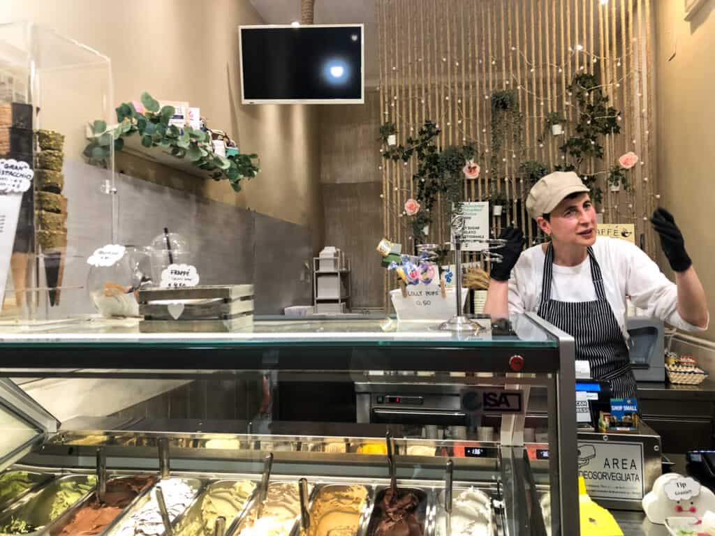 Woman working behind counter at Italian gelateria gestures with both hands. You can see the bins of gelato in the display case in foreground.