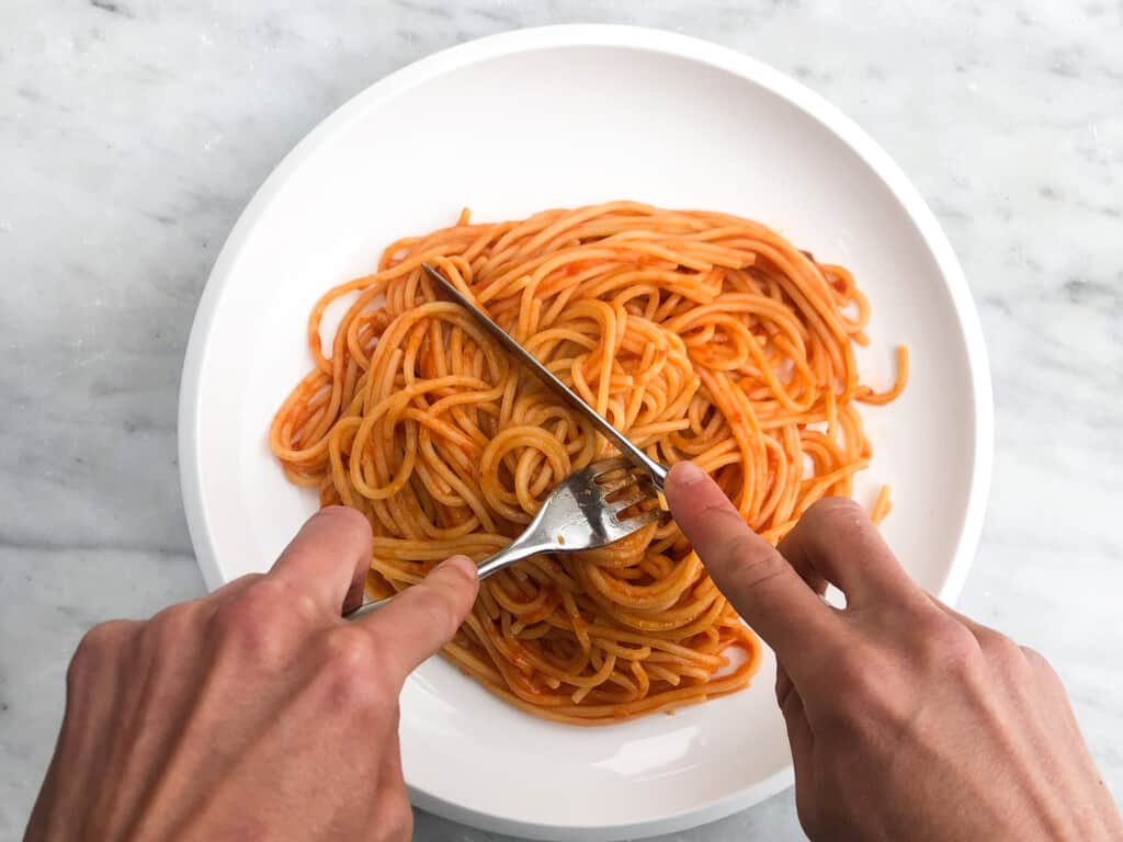 top view of a white plate with pasta with tomato sauce with a pair of hands cutting pasta with a knife and fork