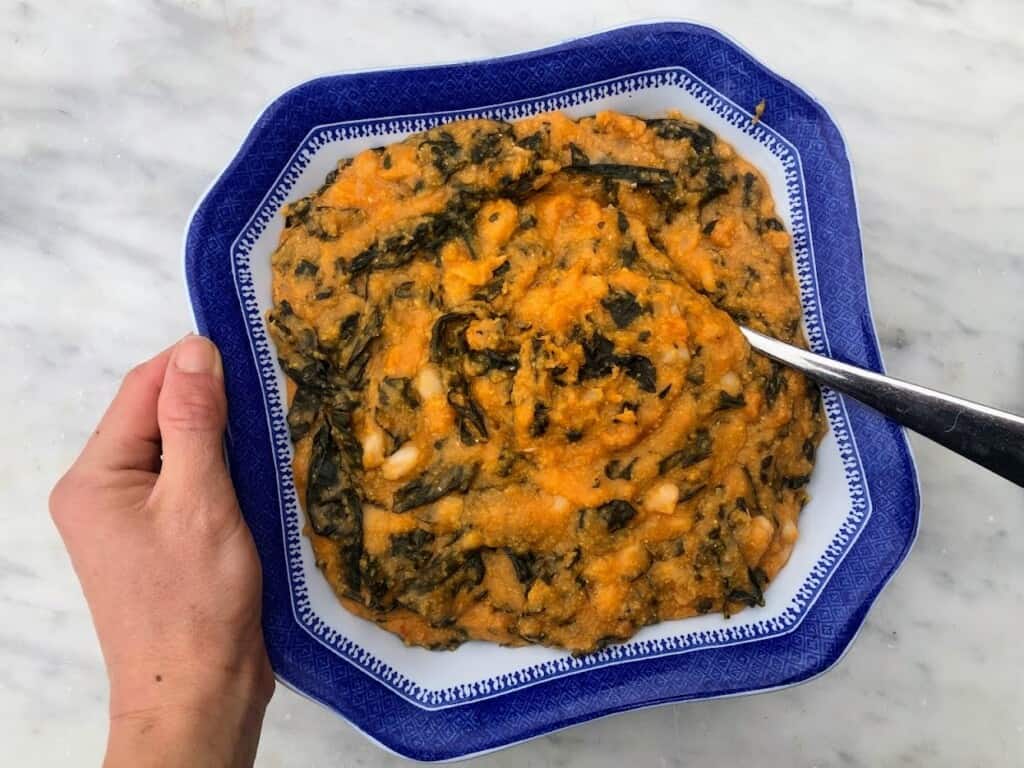 Hand holding blue bowl of farinata con cavolo nero. There is a spoon in the bowl and the bowl is held over a white marble counter.