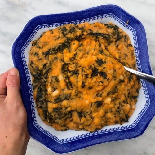 Hand holding blue bowl of farinata con cavolo nero. There is a spoon in the bowl and the bowl is held over a white marble counter.