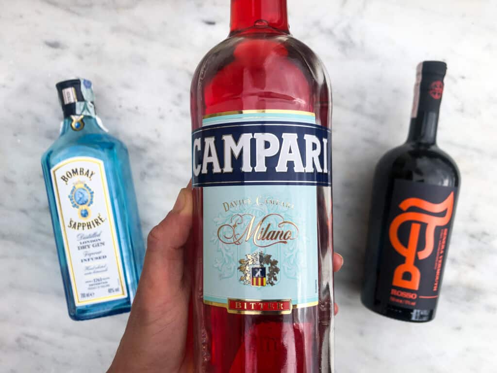 negroni ingredients with gin on left, hand holding campari in middle and vermouth on right on a marble board