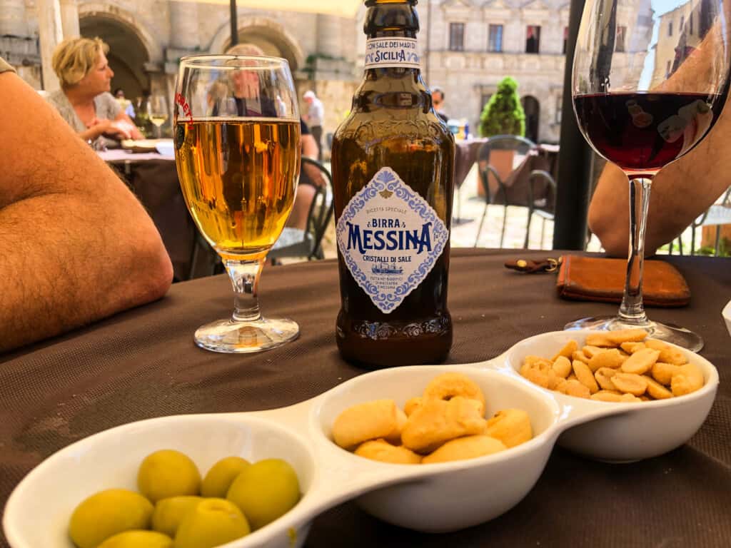 side view of a beer in a glass with a bottle next to it and then in foreground a small tray with nuts, olives and taralli