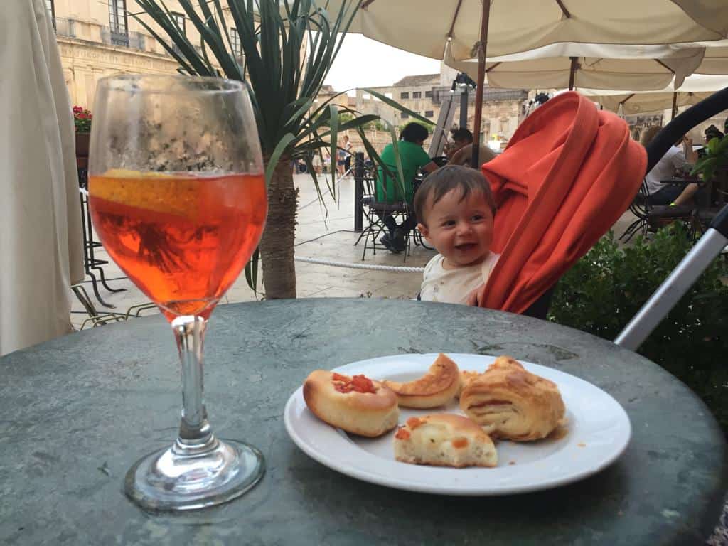 view of a wine glass full of spritz on a table from side view with a small white plate with little appetizers with baby in background at an outdoor bar