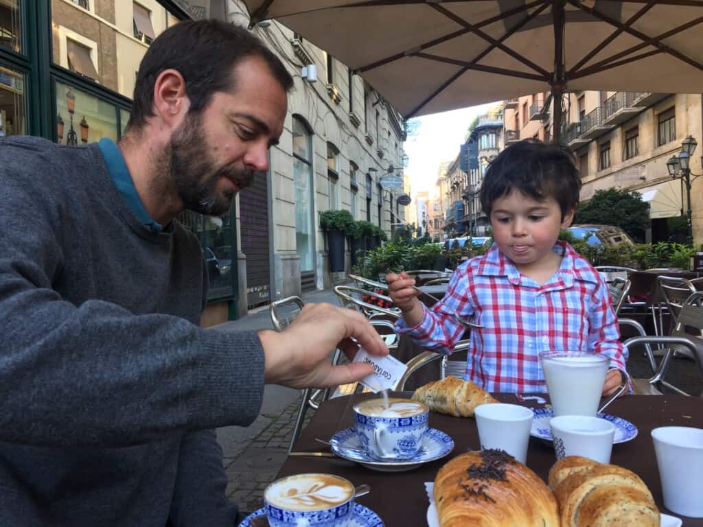 view of a man and a small child at an italian bar outdoors under an umbrella with two cappuccini, several pastries and the man is pouring sugar into the coffee