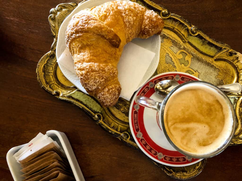 top view of small green tray with a cappuccino and brioche and various decorated pottery on a wooden table with a small dish of sugar packets on the left hand corner