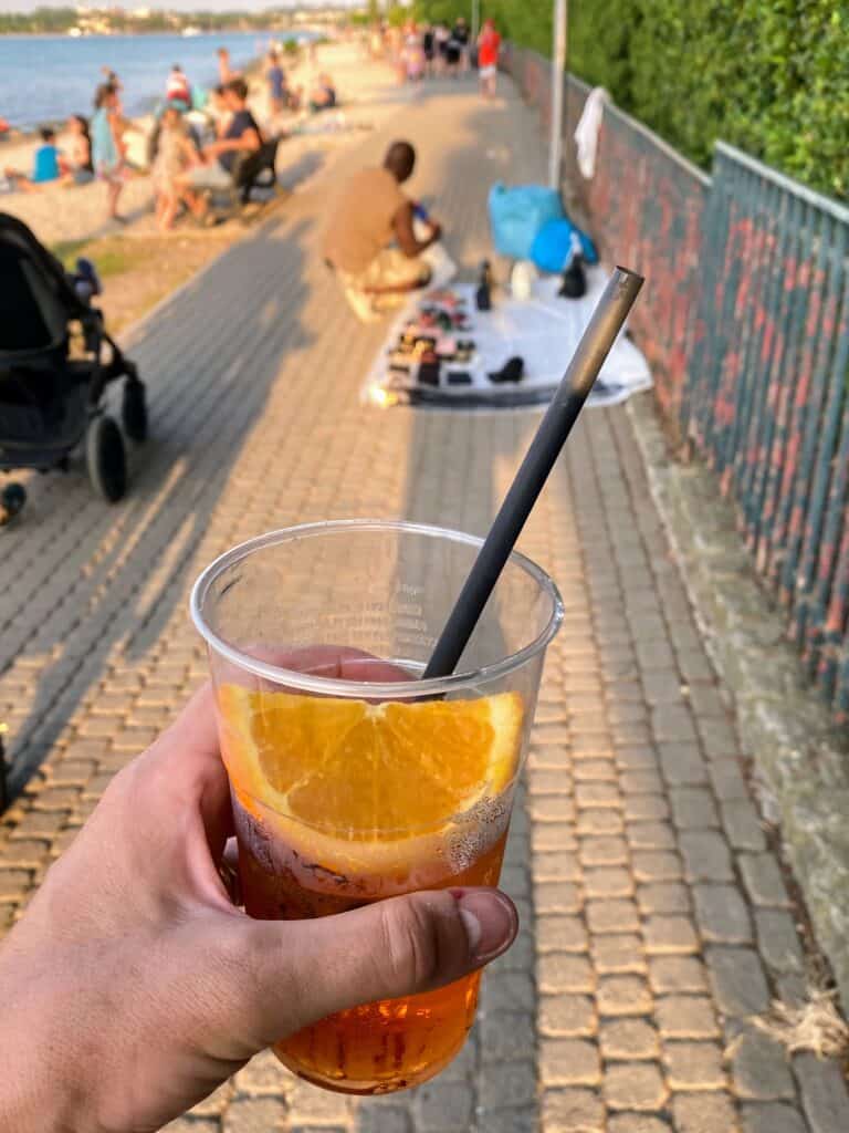 a hand holding a campari spritz in a plastic cup garnished with an orange slice and straw