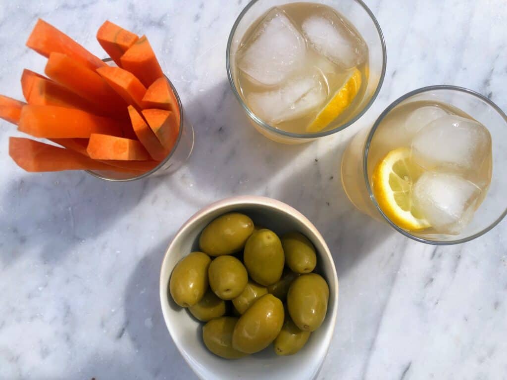 top view of aperitivo with two cocktails, a small bowl of olives and carrot sticks on a marble board