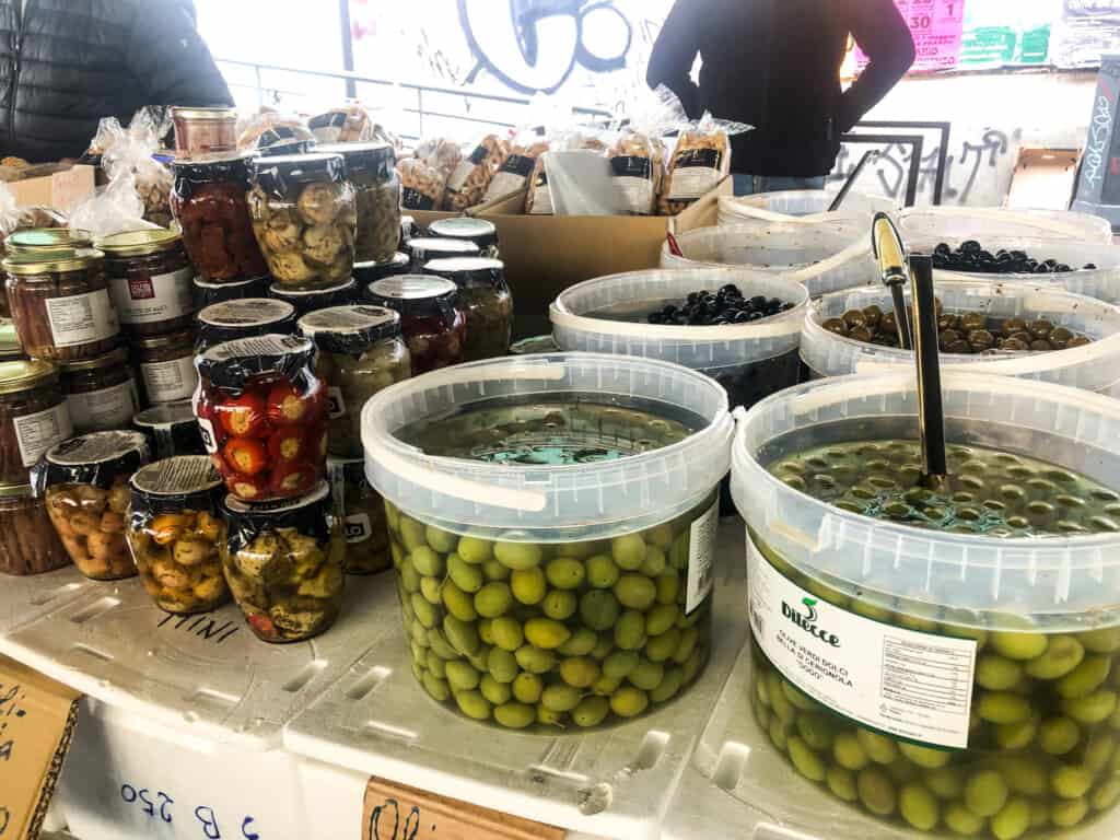 Side view of large tubs of olives for sale with jarred items on side on a table with signs for each item and a man standing in background. 