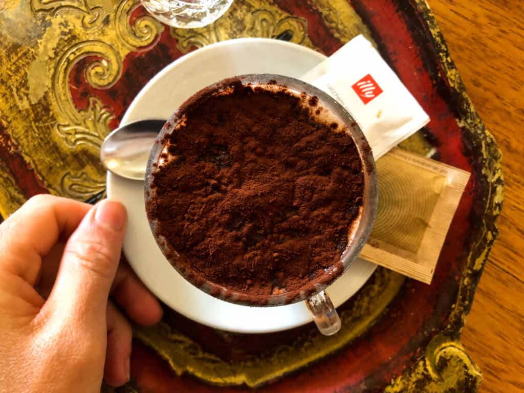 top view of an espressino topped with cocoa powder in a small glass on a white saucer with a hand holding the side on a decorative tray