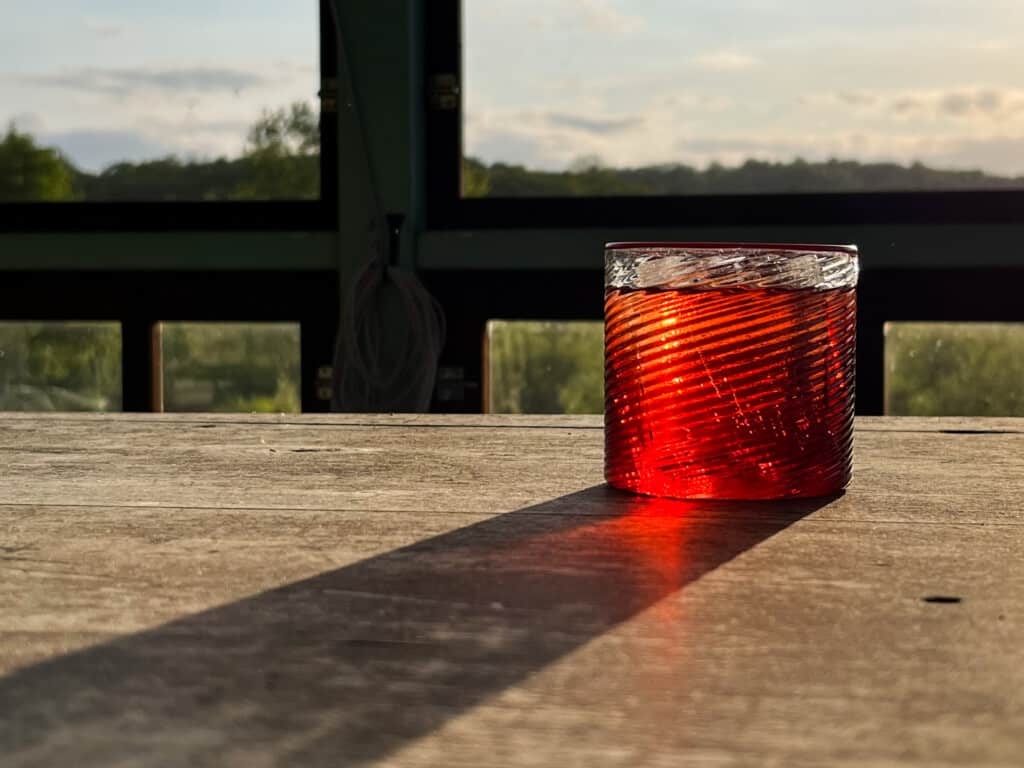 side view of negroni on a wooden table on a porch with greenery in background and shadow of glass coming towards front