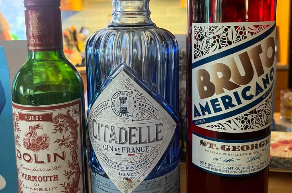 a bottle of brutto american on the far right next to a bottle of citadelle gin in a blue glass bottle and a small green bottle on the left hand side all lined up from side view