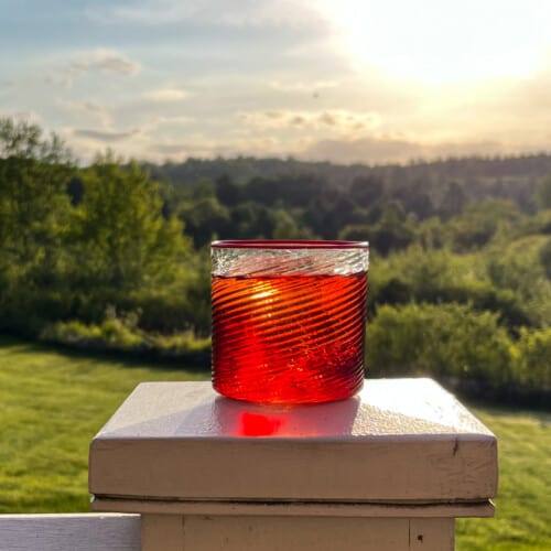 side view of negroni on a wooden table on a porch with greenery in background and sunset