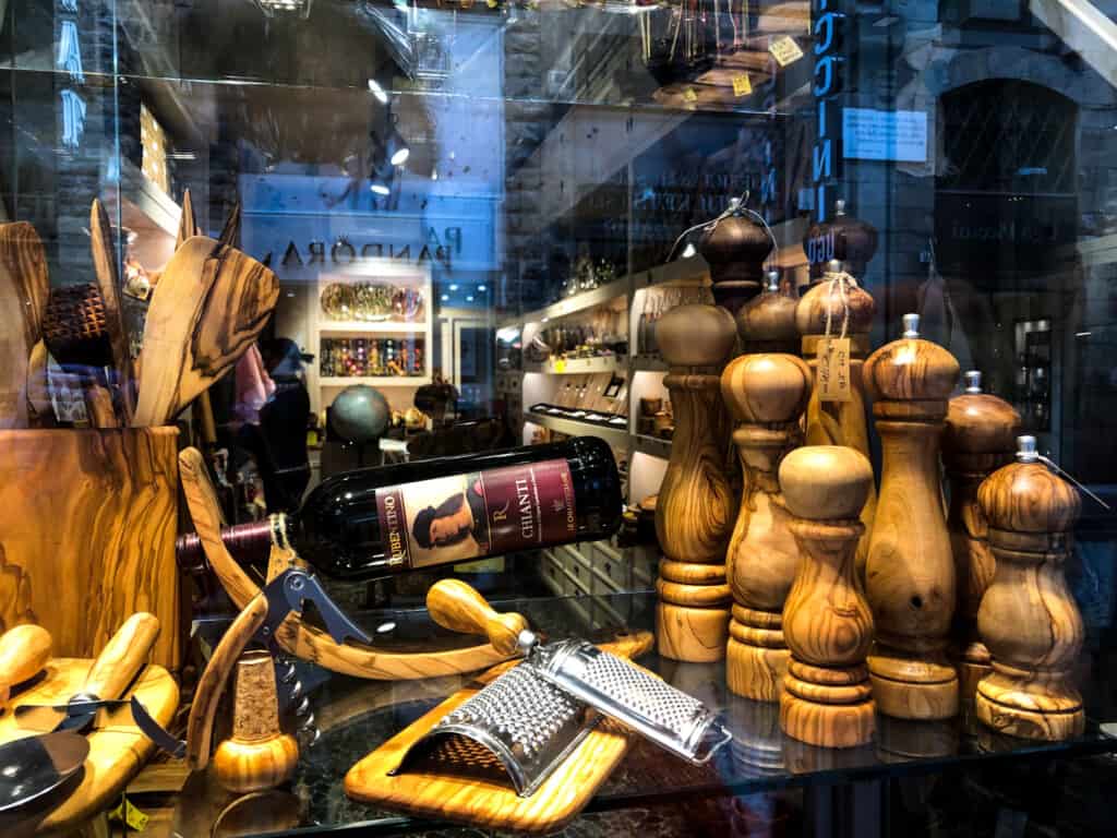 view of a glass storefront window from street with several pepper mills, cheese graters and other wooden kitchen tools all made from olive wood