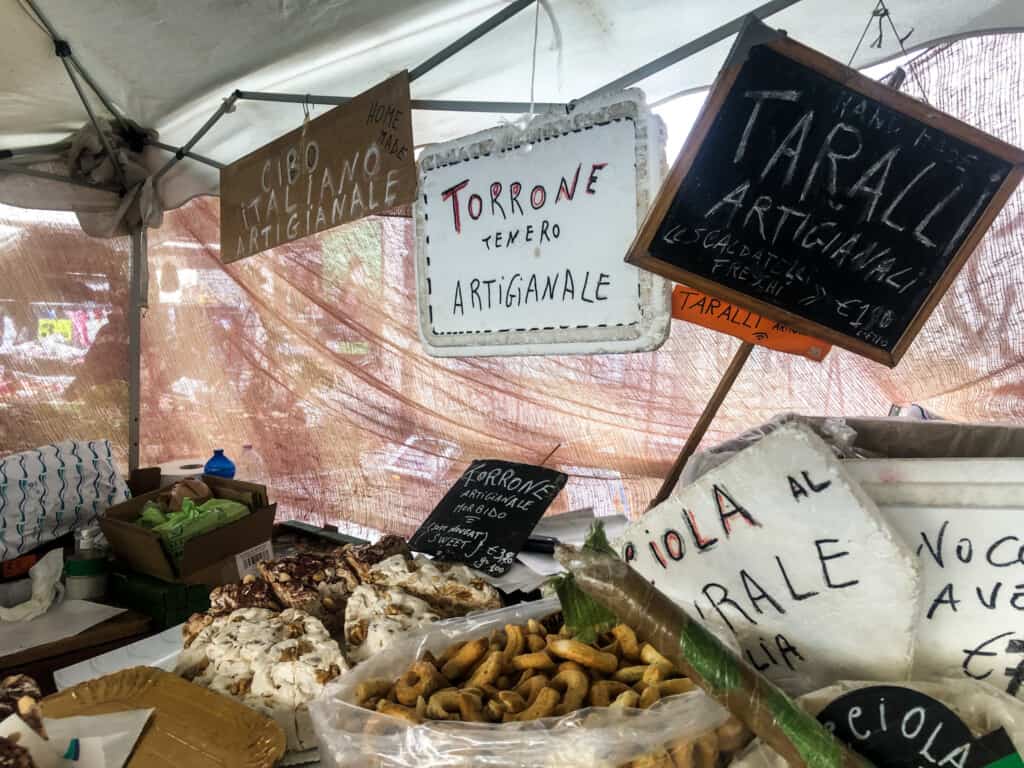 view of a large white sign with written "torrone artigianale" with a big brick of torrone underneath with bags of taralli for sale on side at an outdoor market