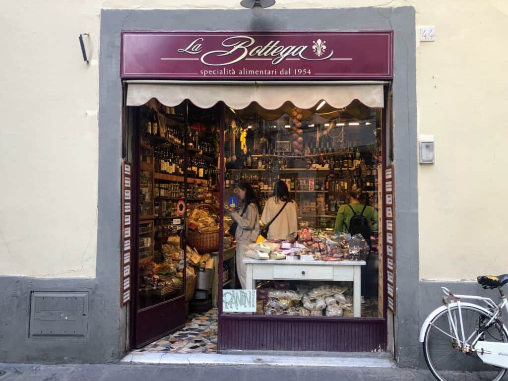 Front entrance of alimentari in Italy. You can see people inside the window shopping for food products in jars, bottles, and small packages.