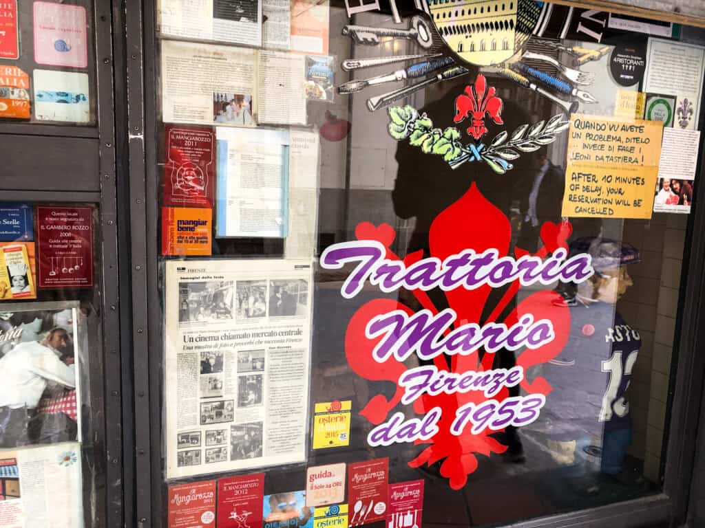 Front entrance window with 'Trattoria Mario Firenze dal 1953' painted in purple. There's also a red giglio painted on the window and newspaper clippings and restaurant award stickers.