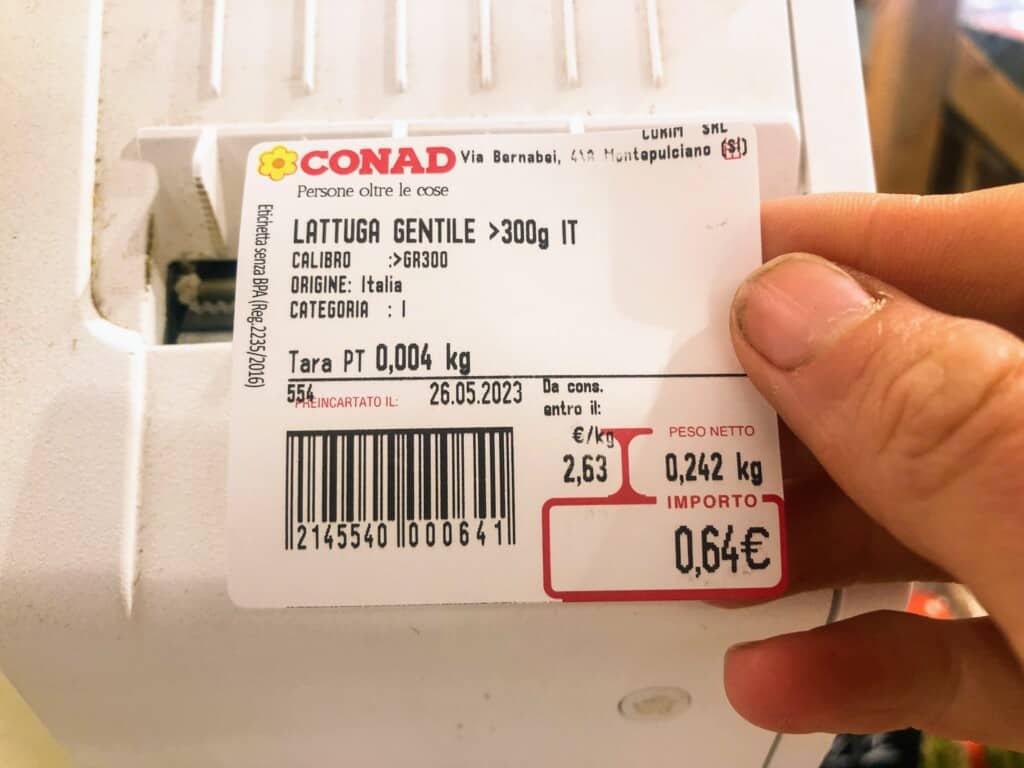 Hand holding price sticker for fresh lettuce in a grocery store in Italy.