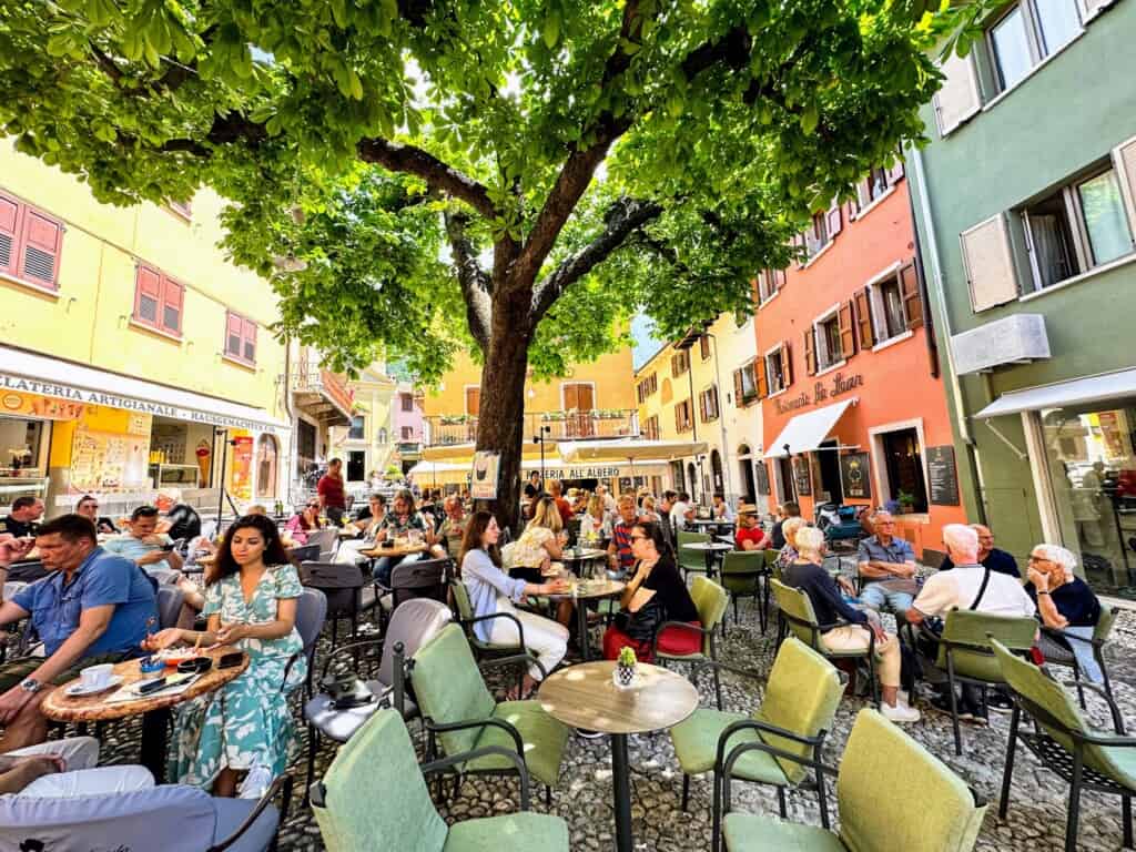 People sitting outdoors in a colorful piazza on Lake Garda. They're drinking coffees and cocktails and chatting.
