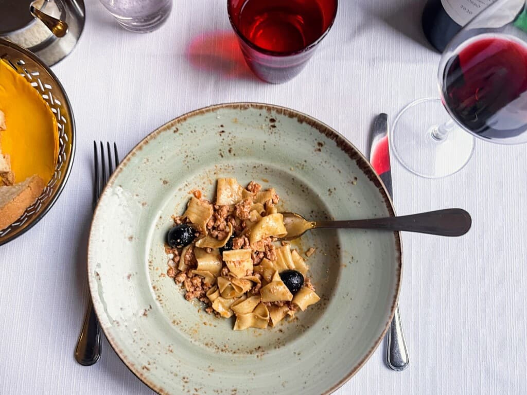 Close up of dish of pappardelle pasta with ragu. You can also see glass of water, glass of wine, and side of bread basket.