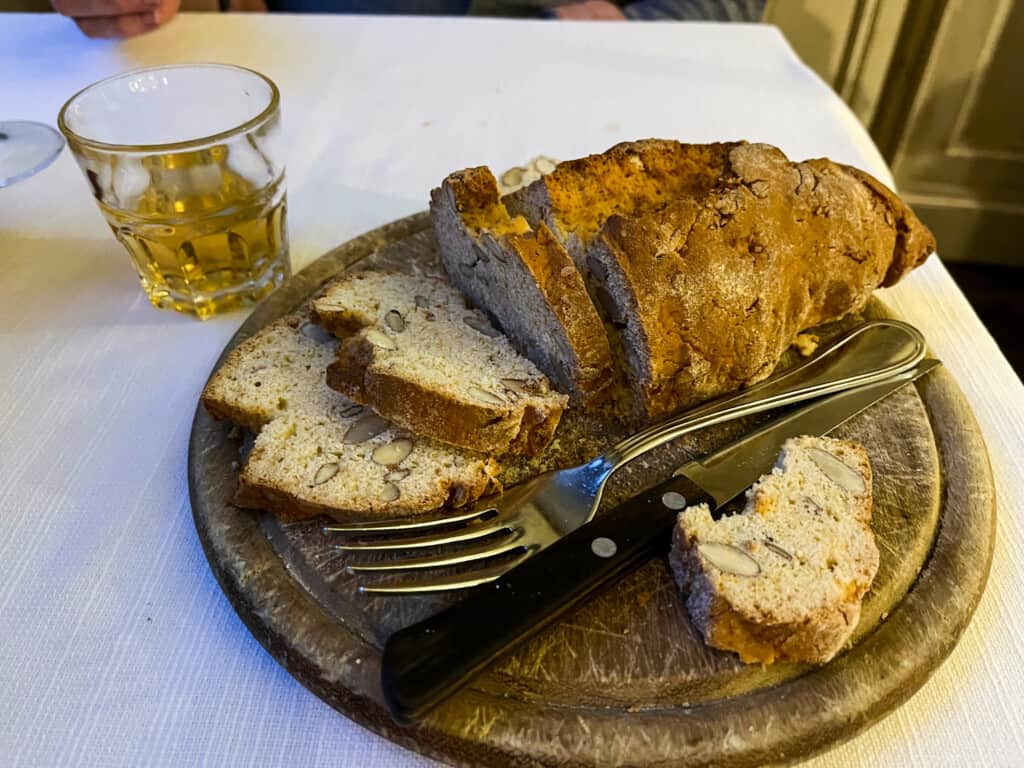 side view of a round cutting board with cantuccini with a fork and a small glass of vin santo to the side on a white table cloth.