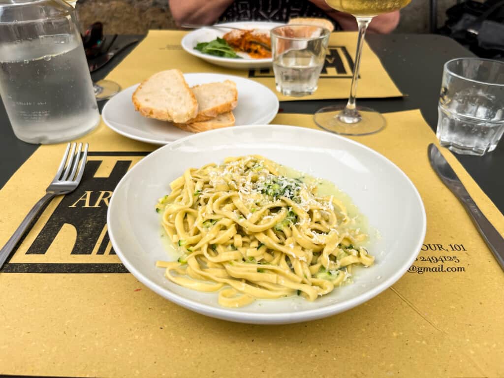 close up of white dish with tagliatelle with zucchini on a table with a yellow paper mat with bread in background on small white plate and glasses with water in background at a table