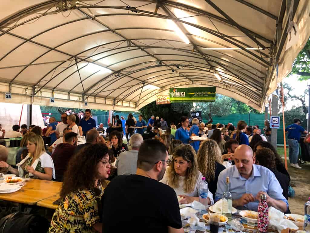 Crowd of people sitting at wooden tables under a canvas roof at a sagra in Italy.