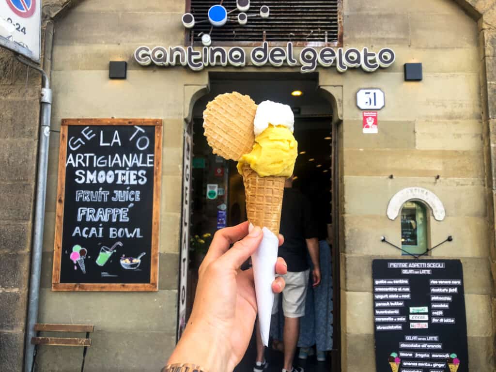 hand holding a large gelato cone with a yellow and white scoop with a cookie on top with a gelateria in background from street view.