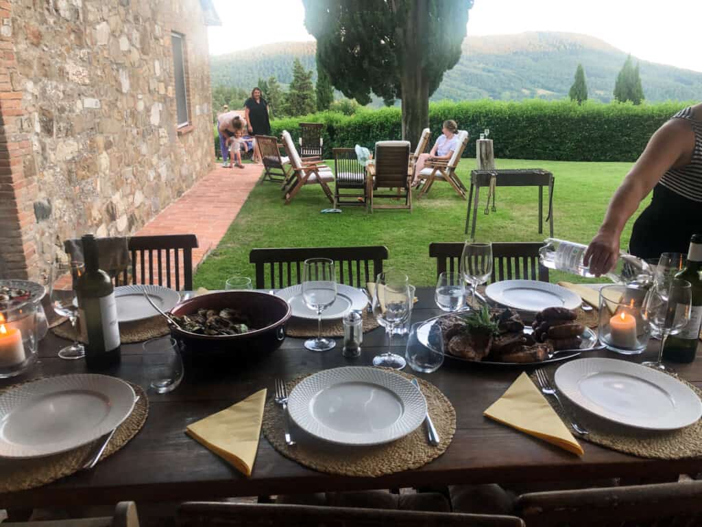 view of dinner table set outdoors on a terrace with green grass and people relaxing in background