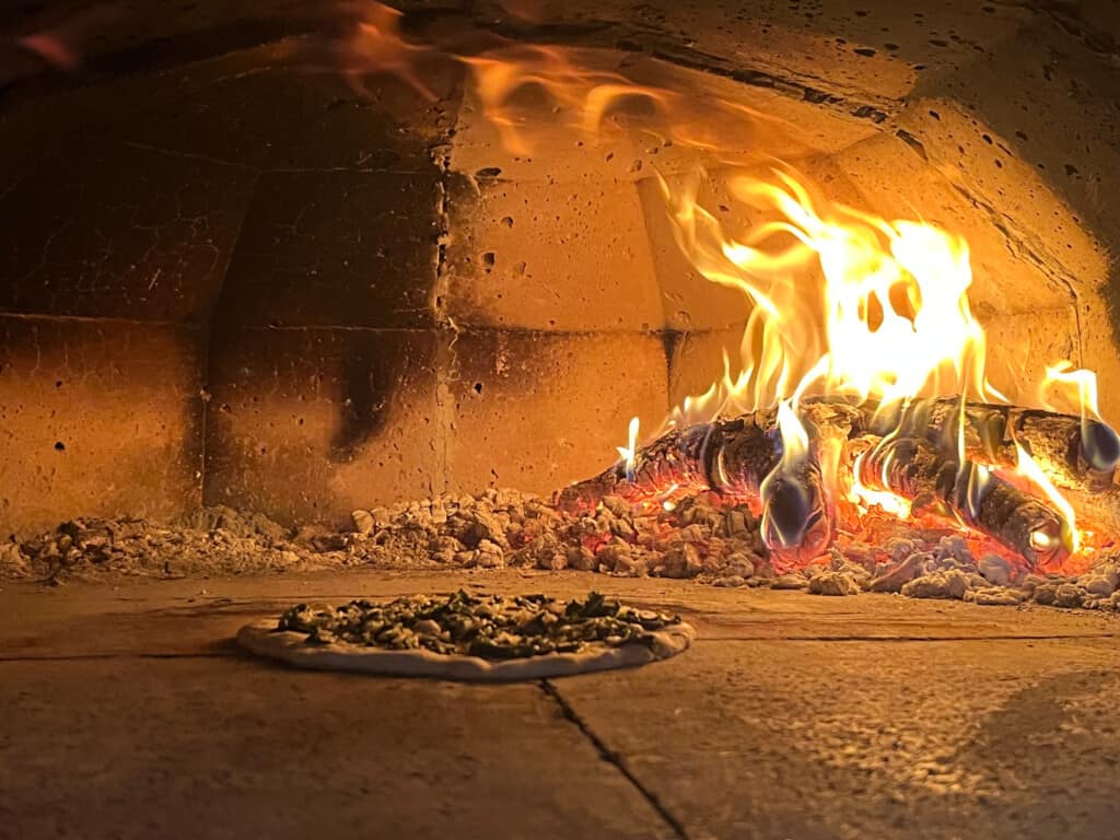one pizzas cooking in a wood-fire oven from side view with big fire flame in back and hard concrete surface in foreground. 