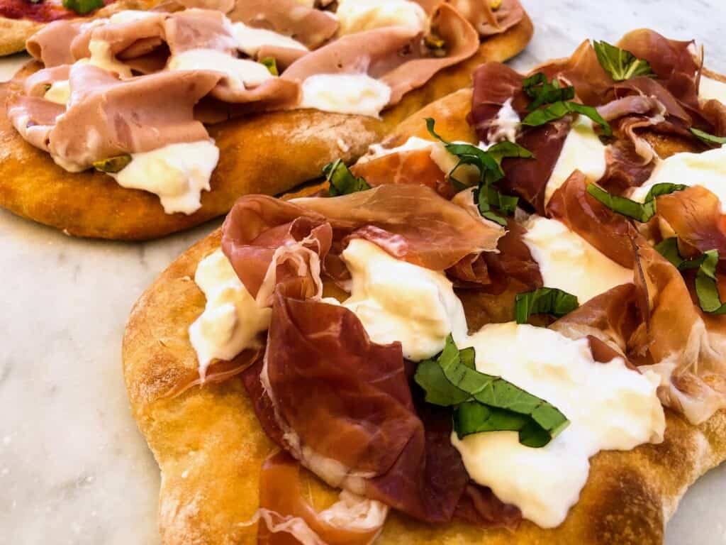 Close up of pinsa from the side. They're topped with proscuitto crudo and cotto and mozzarella and basil.