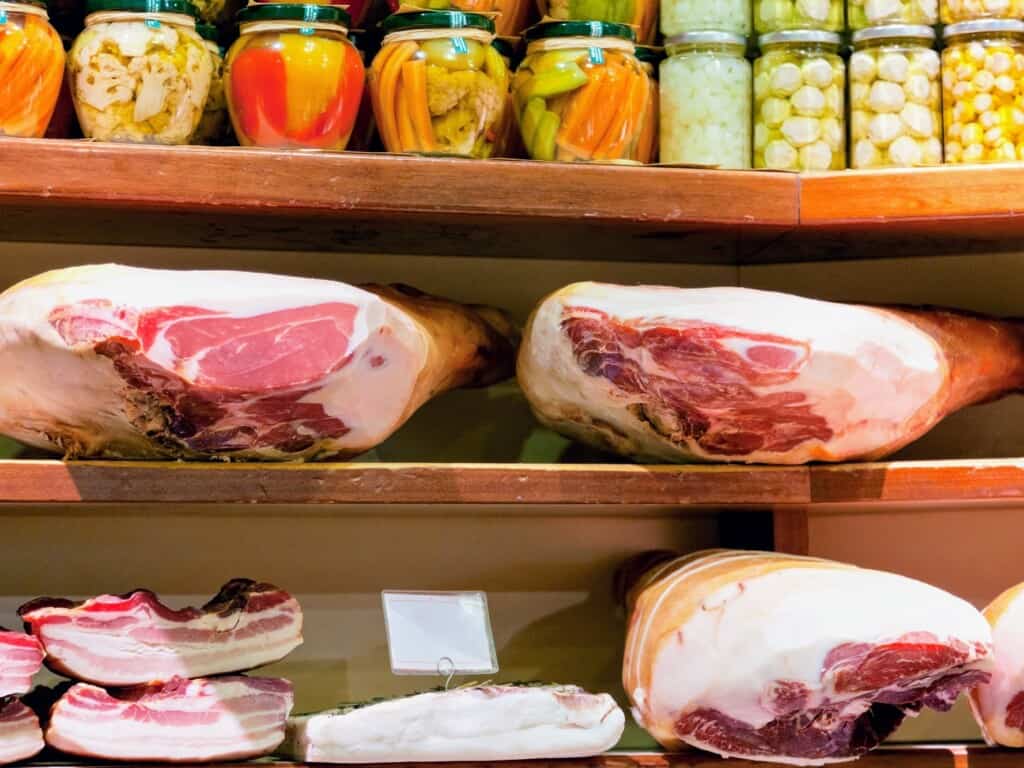 Prosciutto and bacon on display on wooden shelves in a deli in Italy. Jarred vegetables on shelf above.